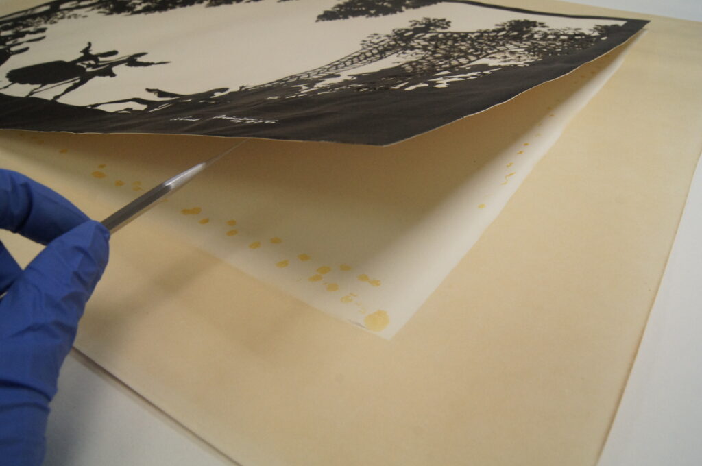 Vanessa's hand using a metal tool to gently lift away the cut-out image from its paper backing