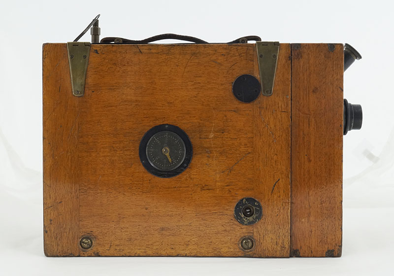 Side view of the camera, which is a simply wooden box with a lens to the front and a carrying handle on top. This side also has a dial.