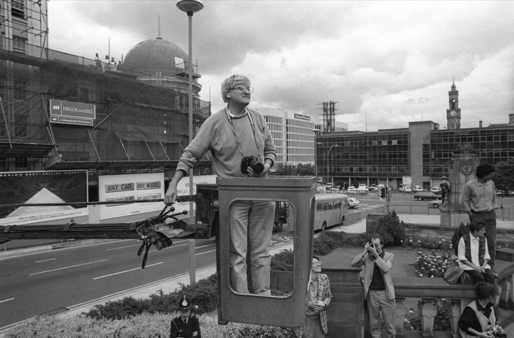 Hockney standing in a cherry-picker by a main road with his camera