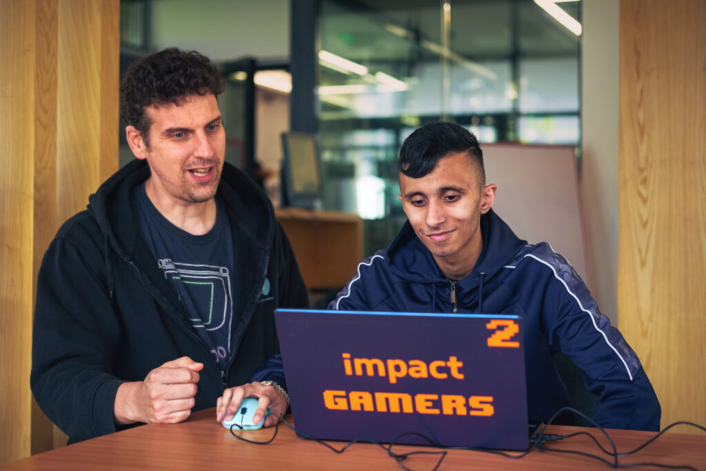 Adam Syrop and a high school student sit at a laptop with the impact gamers logo.