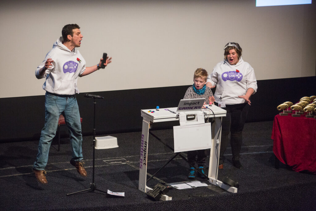 Two people in Impact Gamers sweatshirts jump with excitement as a young child runs a game via a laptop
