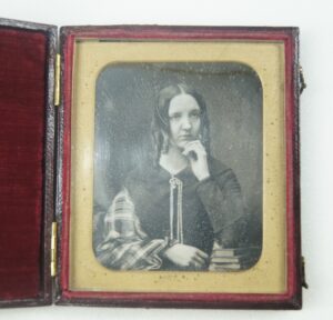 A photographic portrait of a woman with ringlets, resting her chin in her hand. The picture is in a case lined with red velvet, and is speckled with age.
