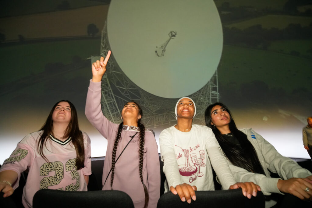 Four students stand looking up, with an image of the Lovell Telescope behind them. One points a finger upwards.