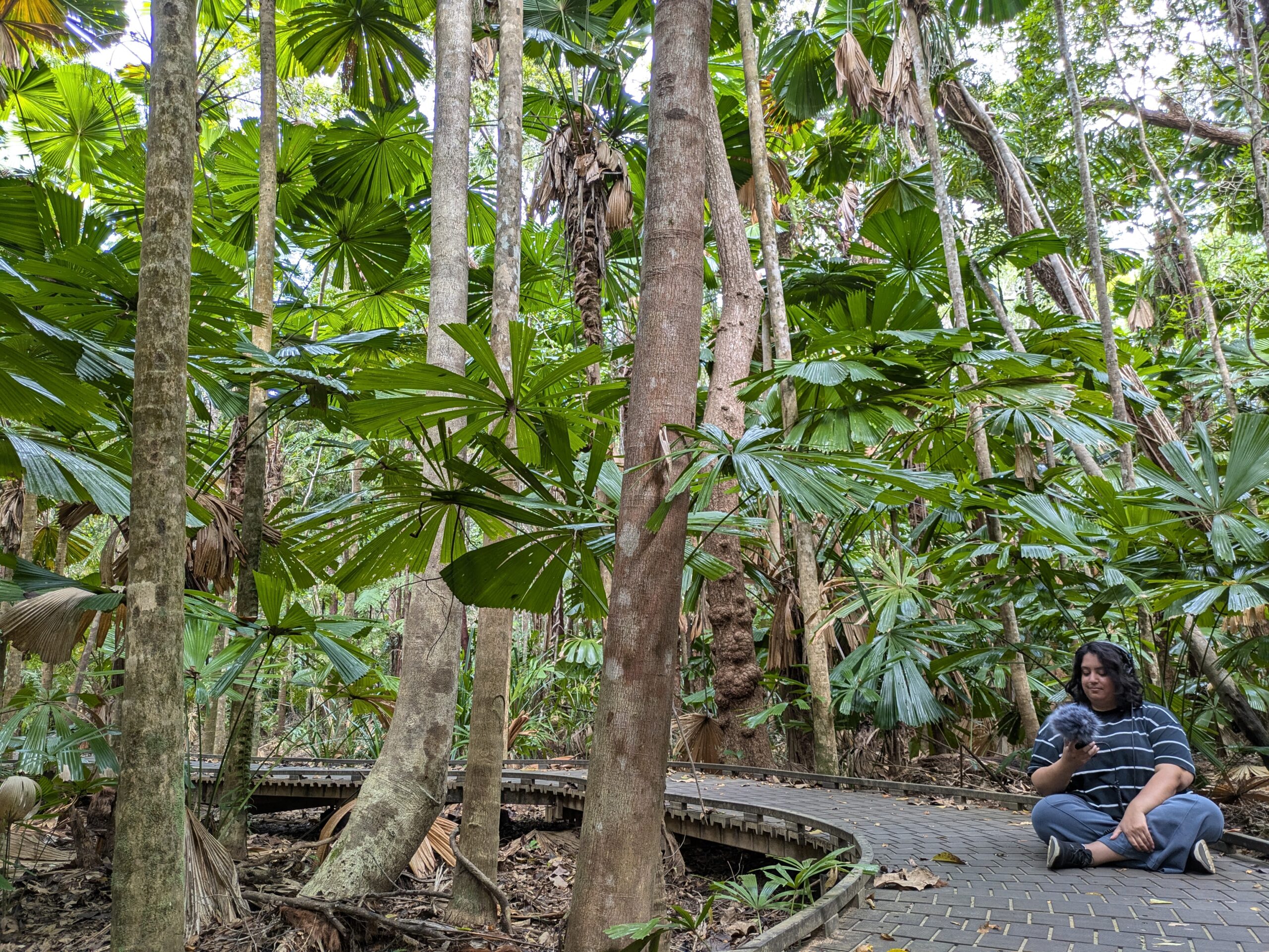 A woman sits cross legged on a path surrounded by large tropical trees. She holds a small microphone with a fluffy boom.
