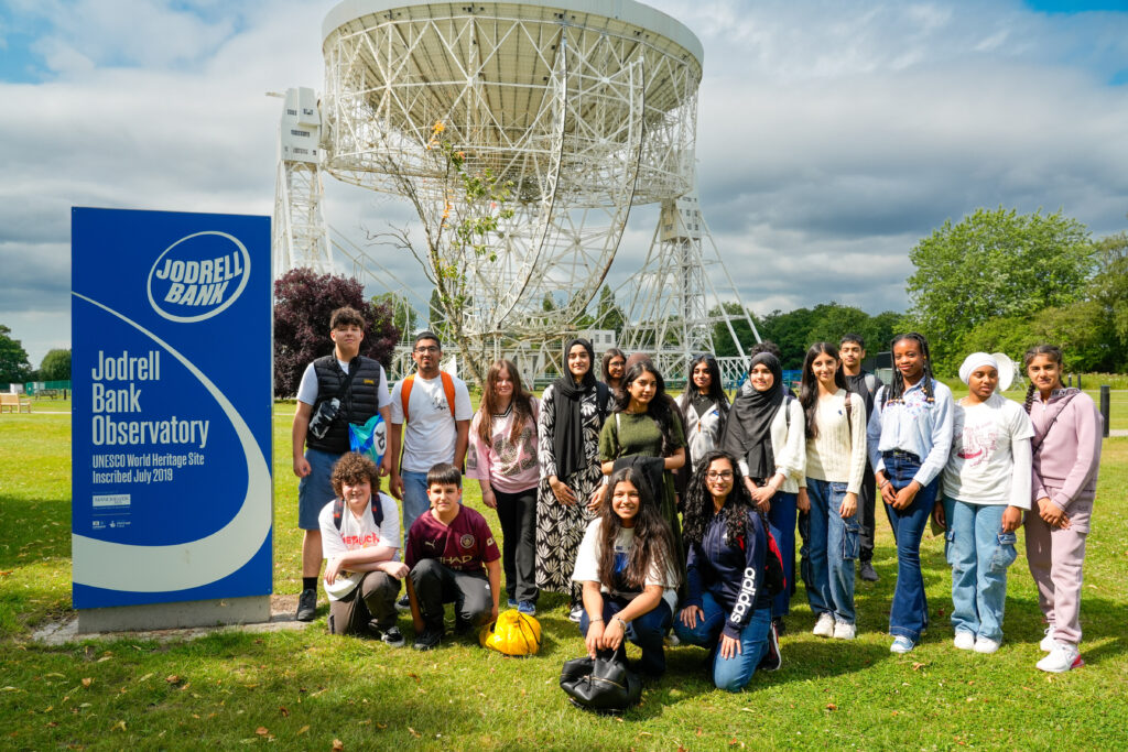 Group of secondary school students standing in front of the Lovell Telescope, a large white half-dome shaped structure, on the lawn at Jodrell Bank observatory.