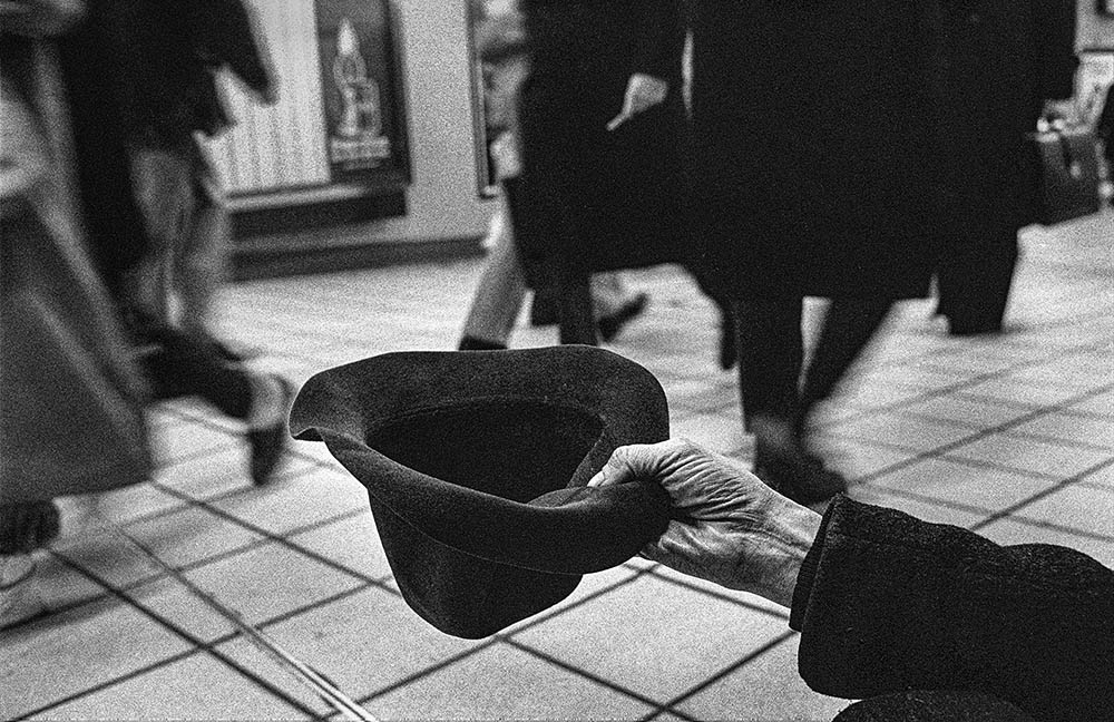 A hand reaches from the bottom right of the frame, holding out a hat as if to ask for change. Behind it, passersby are blurred with motion.