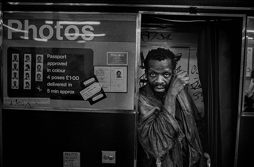 A man in a very worn jacket stands inside a graffitied photobooth, looking at the camera