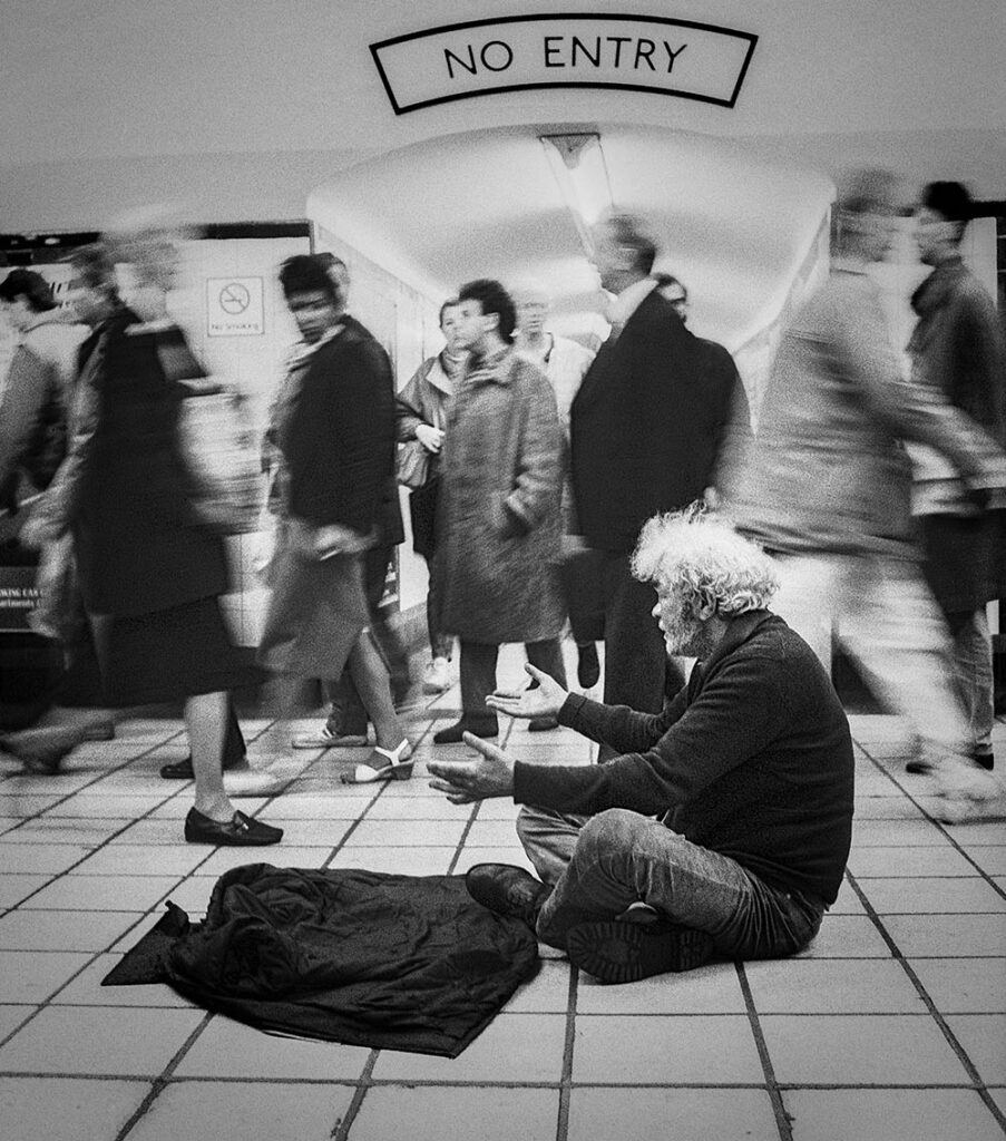 A man sits cross legged on a train station floor with arms outstretched. Behind him, passersby are blurred as if in fast motion.