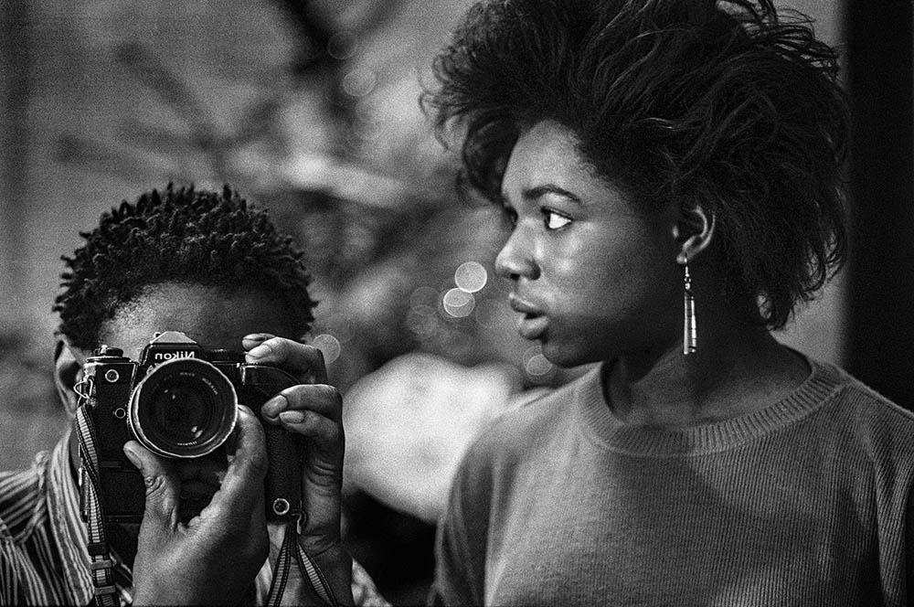 Lanre Fentinola holds up an SLR camera. Next to him is a woman with large silver earrings, who looks over his shoulder out of shot.