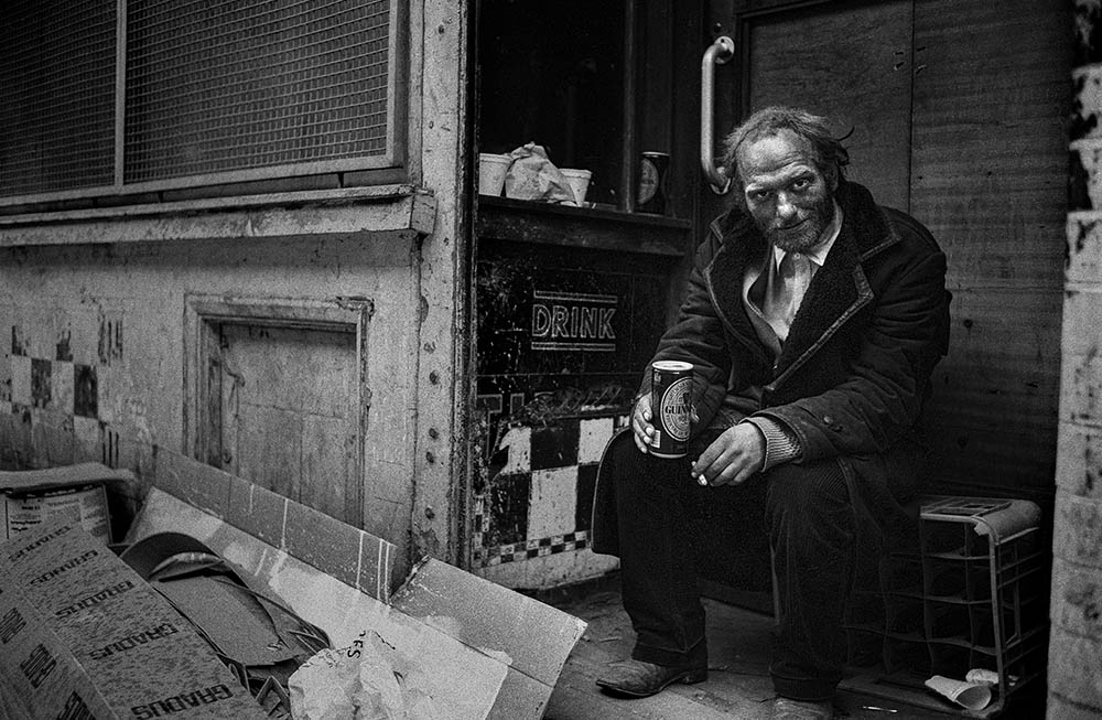 A man wearing a leather overcoat sits smoking and drinking a can of Guiness in a shabby corner filled with waste carboard