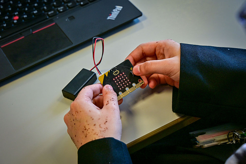 A young person's hands holding a microbit - a small board with a grid of red lights attached by short wire to a battery