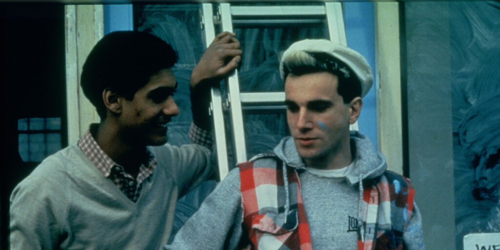 Johnny and Omar lean against a ladder outside the laundrette. Johnny wears a white cap, grey hoodie and red plaid overshirt. Omar wears a grey sweater and collared shirt.