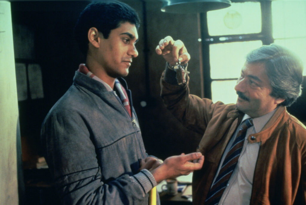 Nasser holds a set of keys out to Omar, who holds his hand out for them. Nasser has a knowing smile, Omar looks serious.