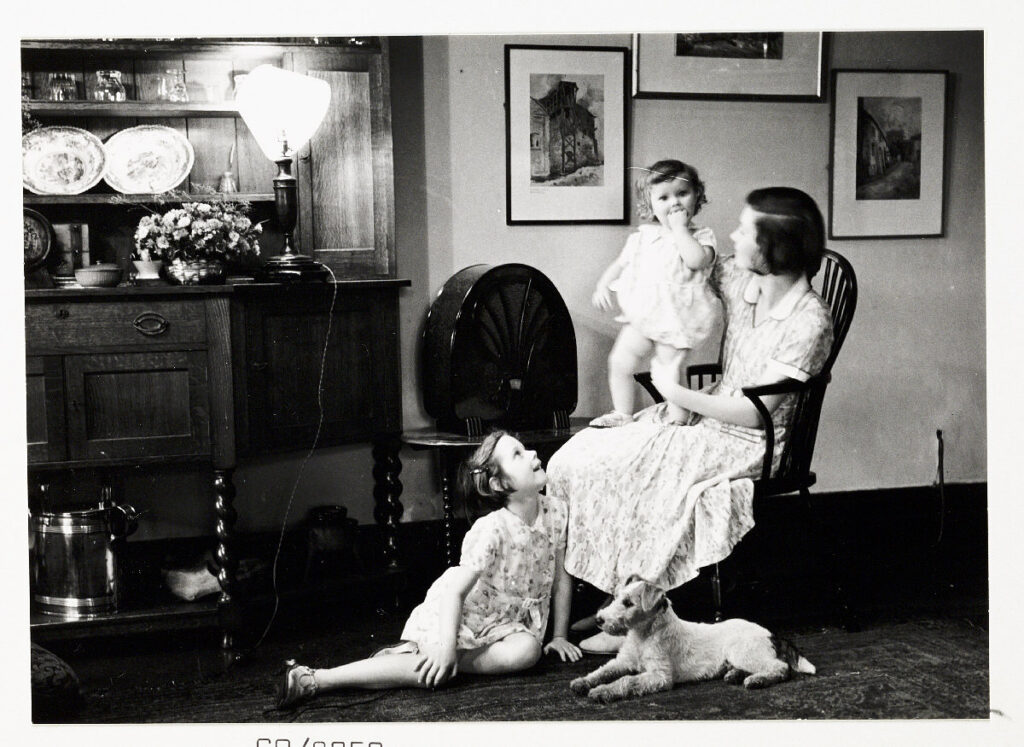 A woman sits by a wireless with a baby on her knee, and a young girl and a dog sat at her feet,