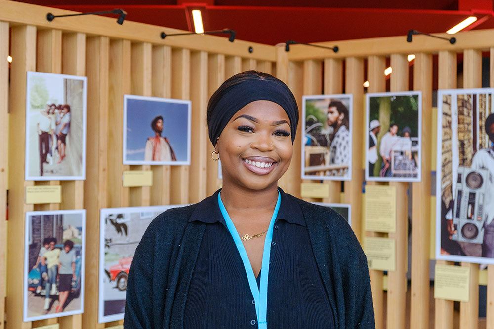 Leah, a young Black woman wearing a black top and headband, smiles in front of a photography display