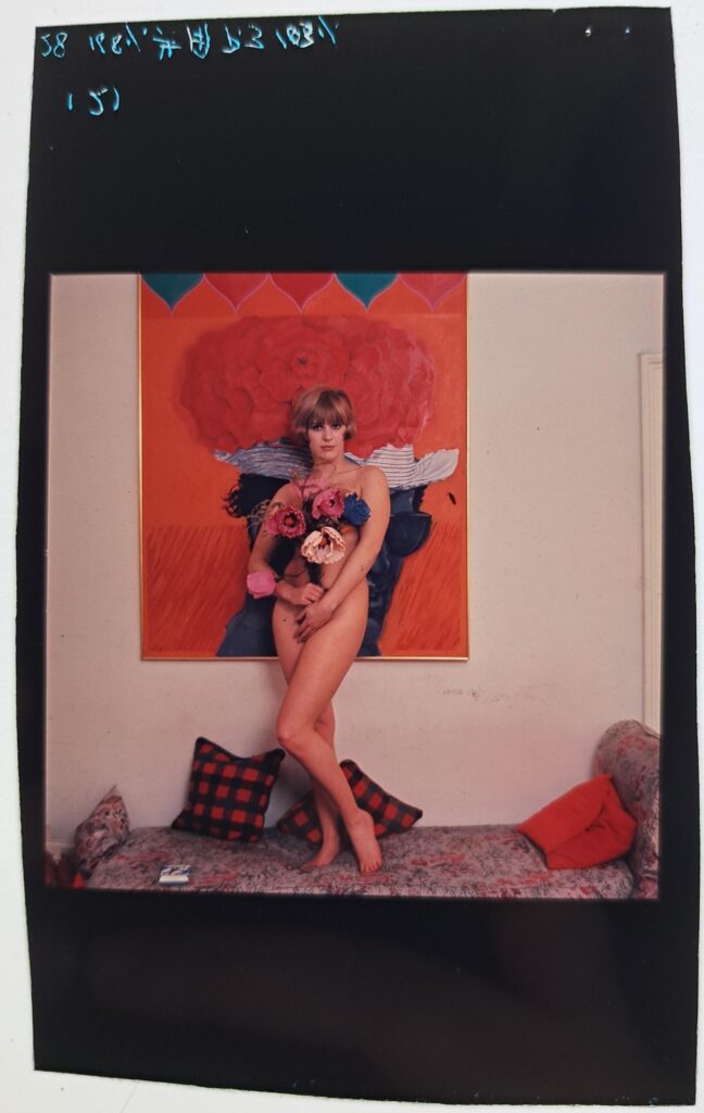 Pauline stands nude on a floral settee holding a large bunch of flowers, with her brightly coloured painting on the wall behind.