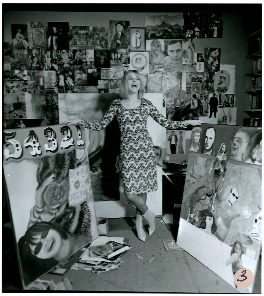 Pauline wears a patterned dress and a joyful expression, as she stands holding two large paintings up. The wall behind her is covered with photographs.