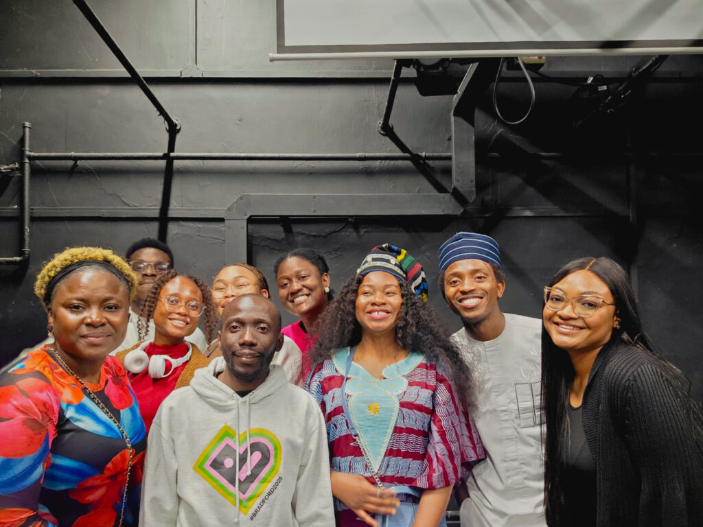 A group of Black creatives stand against a black studio wall and smile at the camera