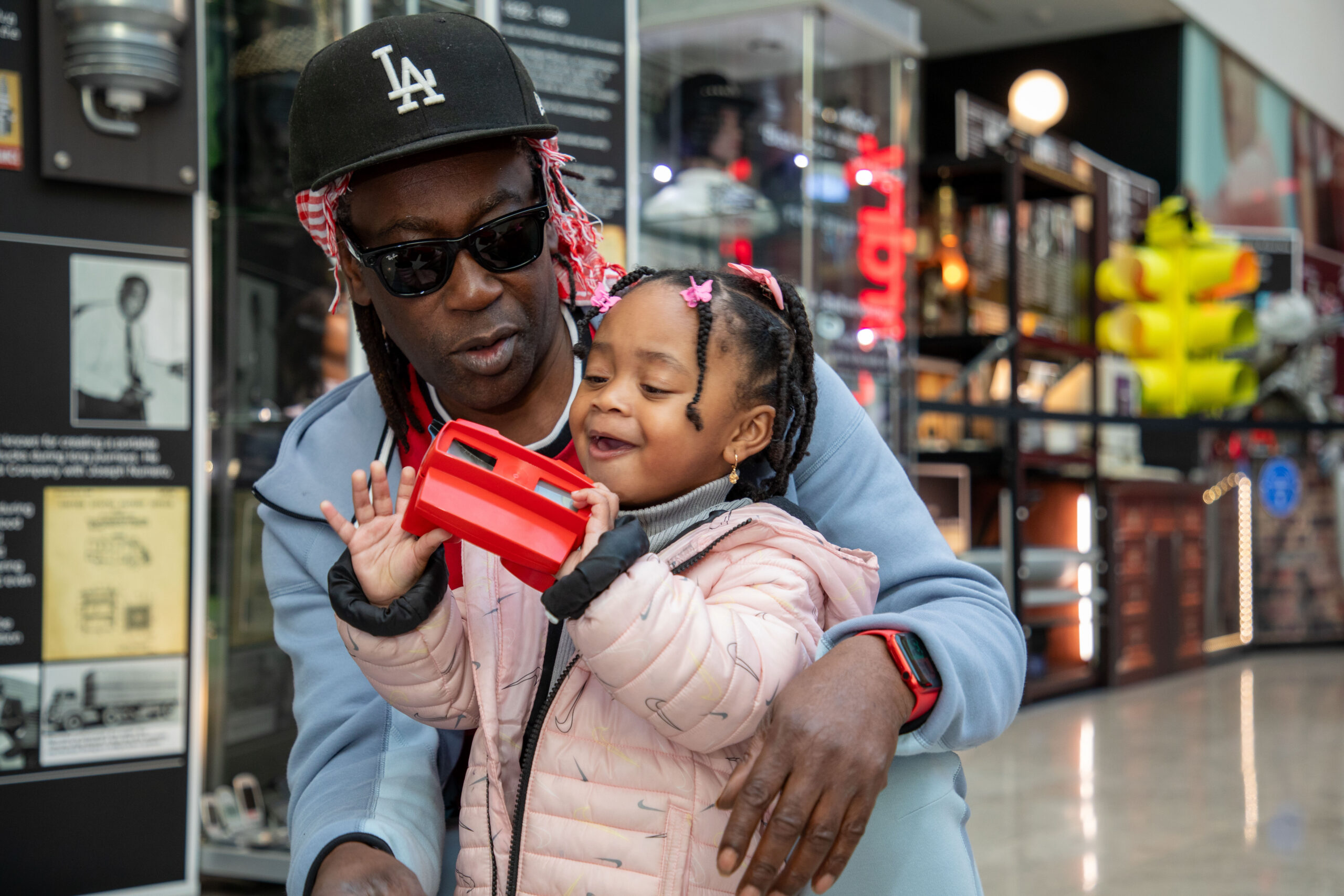 A man in sunglasses and a small girl in front of the Real McCoy exhibition. The girl smiles and plays with a red plastic image viewer.