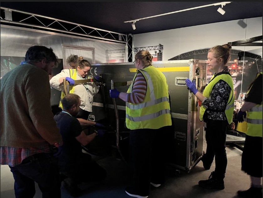 A large black case being tilted by a group staff, some of whom wear yellow hi vis