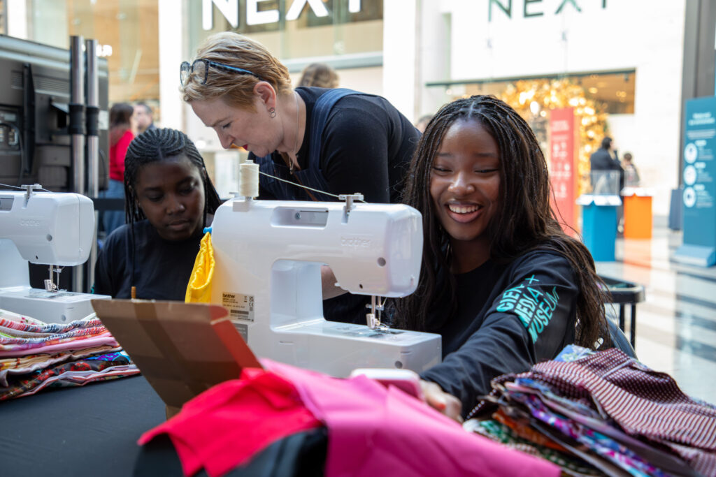 Two young women taking part in a sewing activity with sewing machines and colourful fabrics. An older woman helps them.