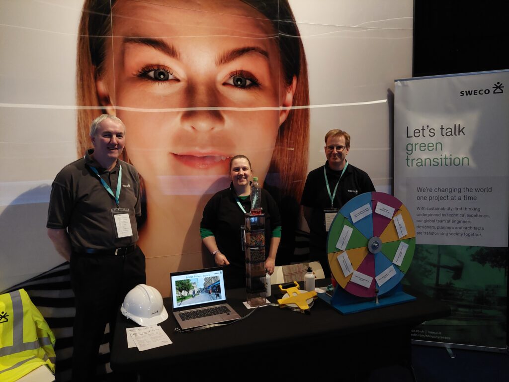 Three adults stand by a table with information about working as an engineer. Behind them is a large image of a woman's face.
