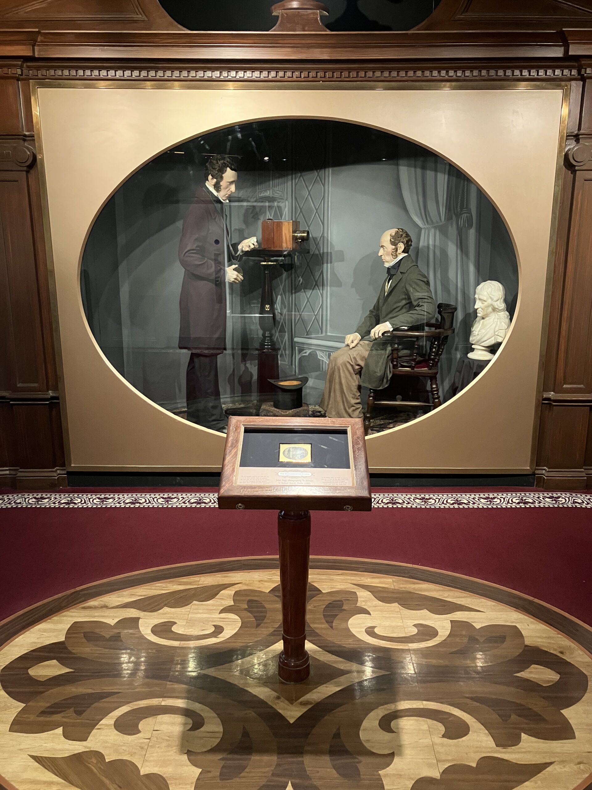 A glass museum case with dummies of a Victorian photographer and his sitter.In front of the case is a small pedestal with a framed photograph of the scene.