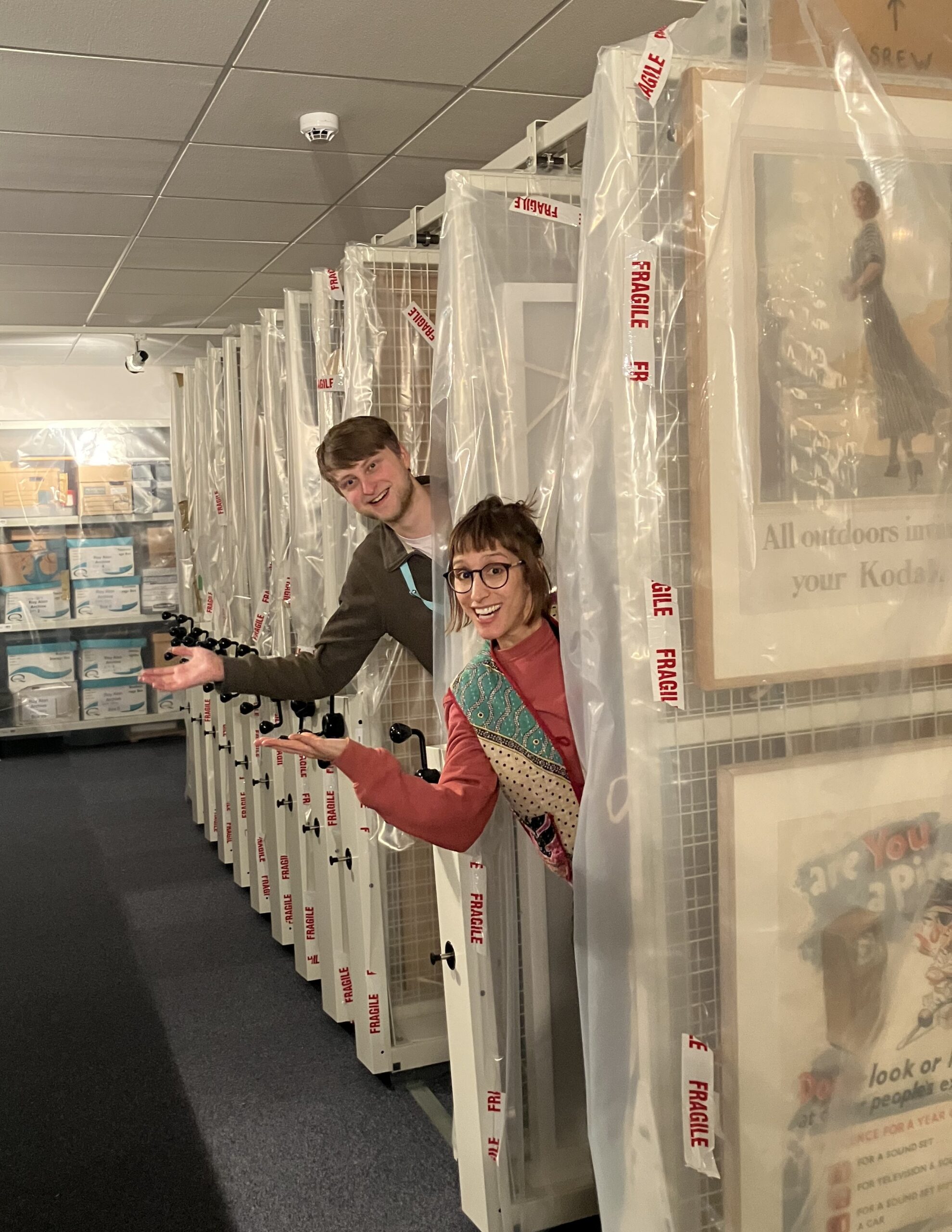 Vanessa and Ben lean out from between racks in a frame store, doing jokey chorus line poses
