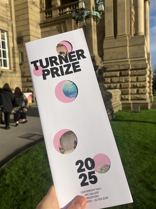 A hand holds up a leaflet for the Turner Prize 2025 in front of Cartwright Hall art gallery.