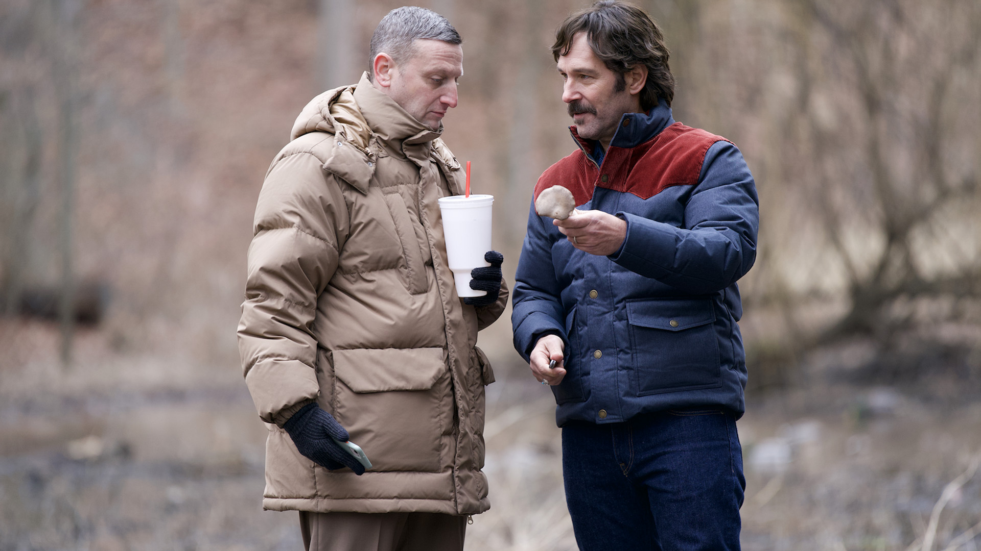 Two men in warm jackets stand in a winter wood. One holds a mushroom and the other a large takeaway drink cup.