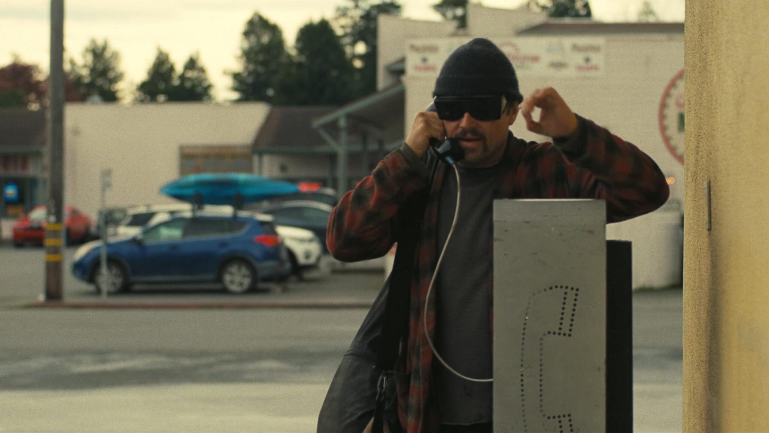 A man wearing dark glasses and a beanie hat talks on a public payphone in a car park.