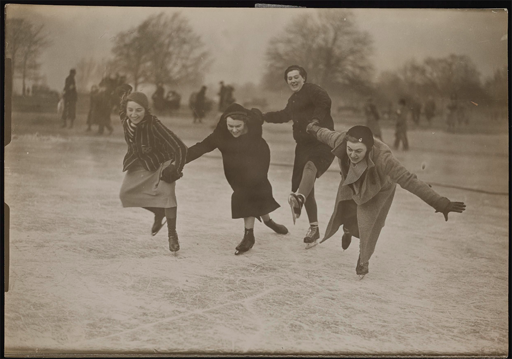 Four women hold hands, laugh and lunge wildly as they try to remain upright while skating on a frozen lake.