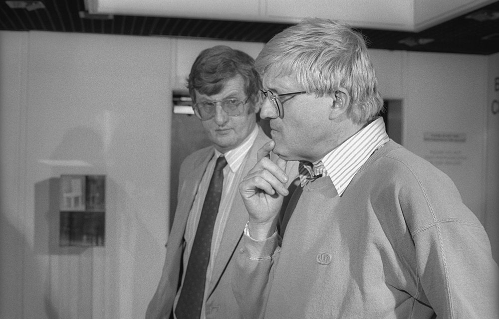 Colin Ford stands next to David Hockney, who holds his hand to his chin pensively.