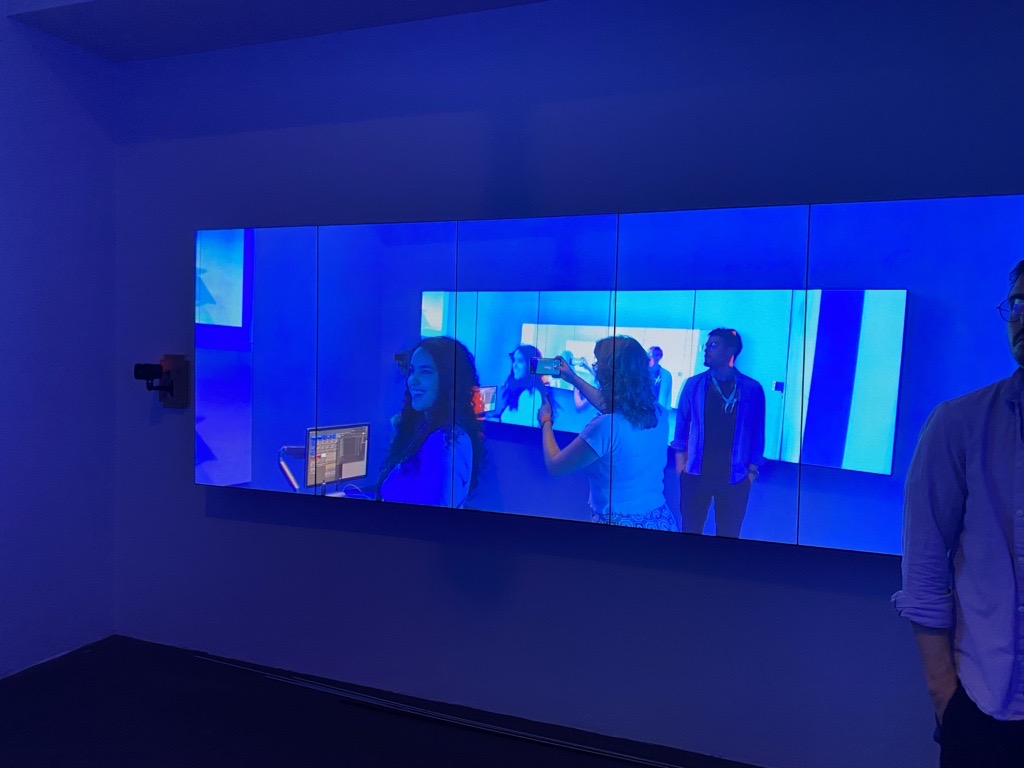 A blue video screen on a wall shows Iman smiling alongside two other people, the image reflected repeatedly behind her.