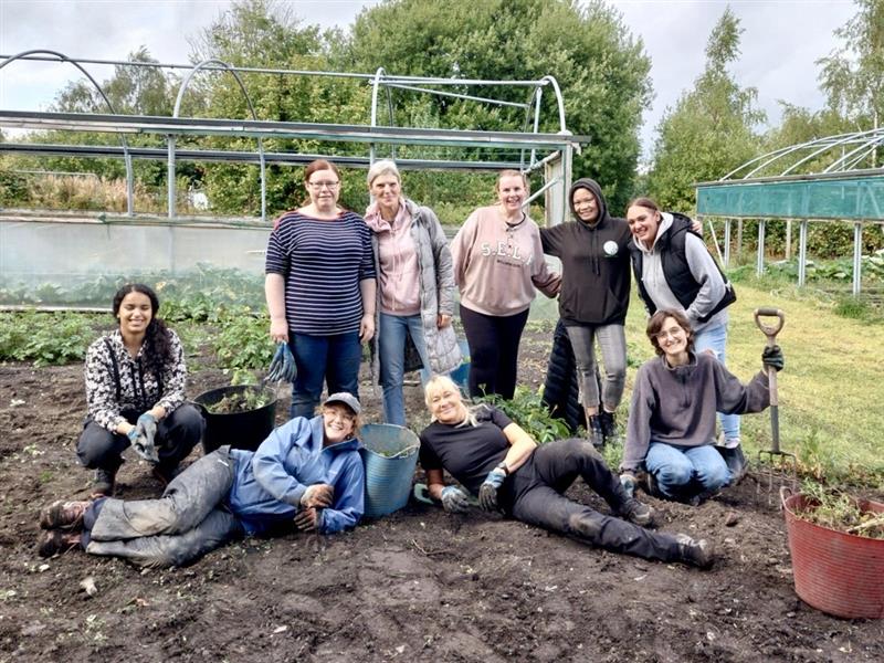 A group of people stand and lie on muddy ground in front of polytunnel frames and trees. Iman crouches on the left and everyone smiles broadly.