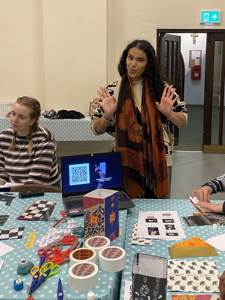 Iman gives a cheeky smile and holds up her hands in front of a table laid with craft materials. Another person sits next to her.
