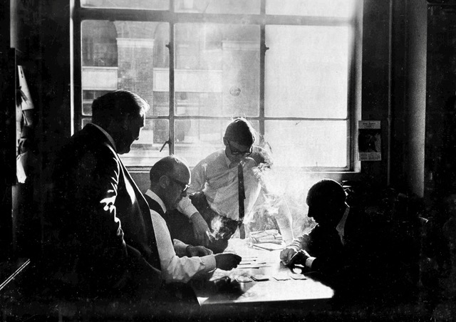 Four men sit at a table, backlit by the large window behind them and surrounded by cigarette smoke.