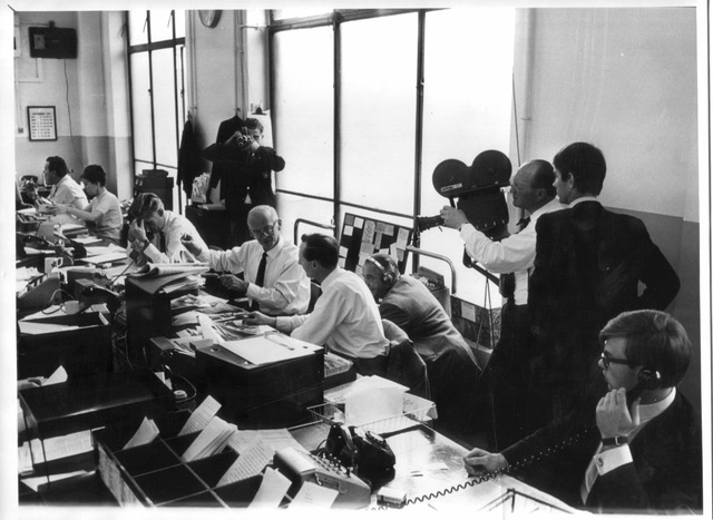 A long row of crowded desks cluttered with files and papework. A cine camera operator and photographer record the scene.