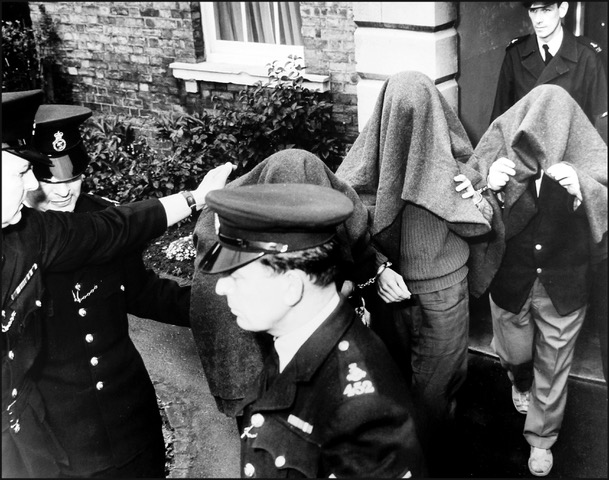 Three men with coats over their heads leave a house, surrounded by uniformed police officers.