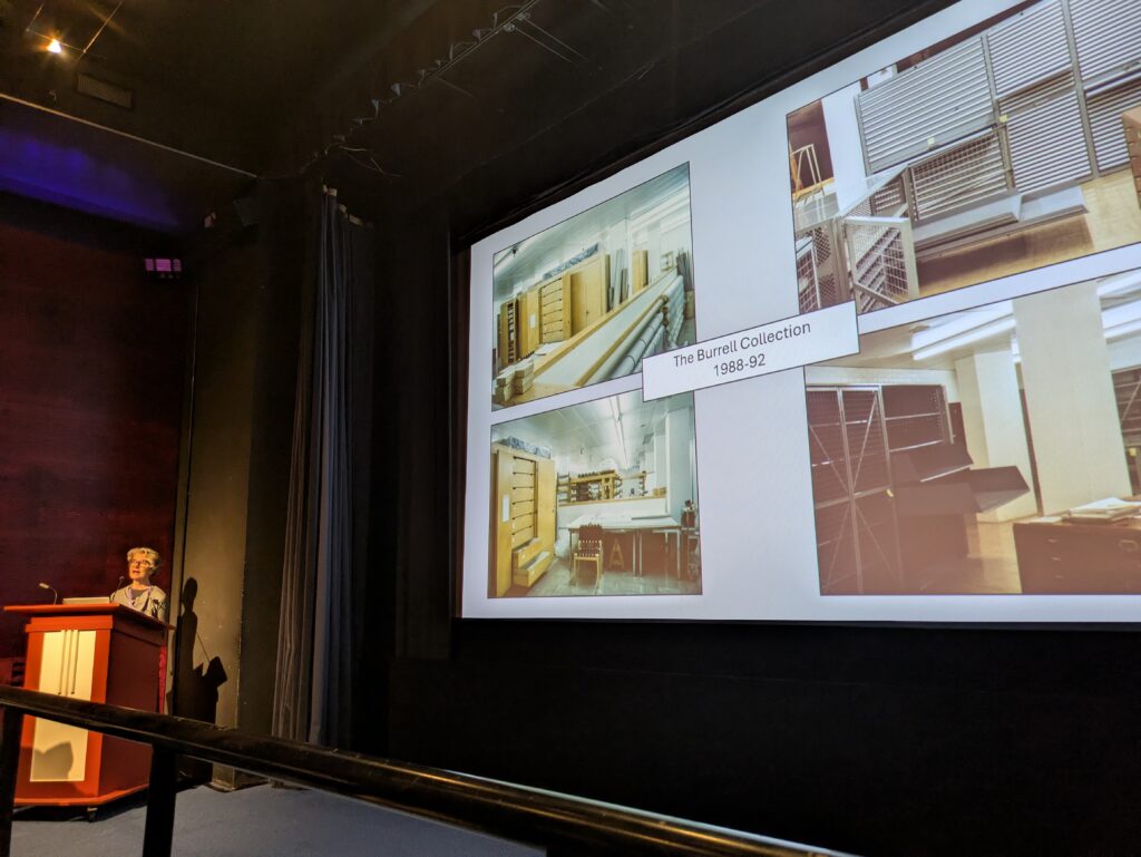 Ann speaks from the lectern in the cinema, with a slide on screen showing four photos of stores titled 'the Burrell Collection 1988-92'