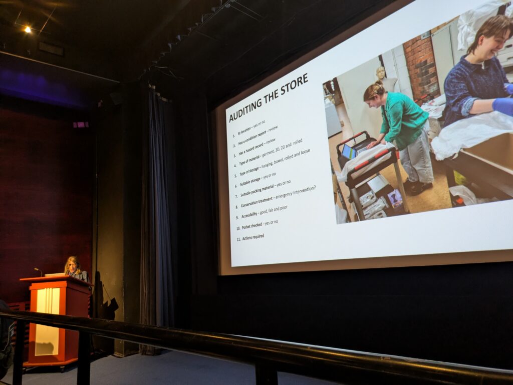 Kathryn gives a presentation from the lectern in the cinema, with a slide of text and an image title 'auditing the store'