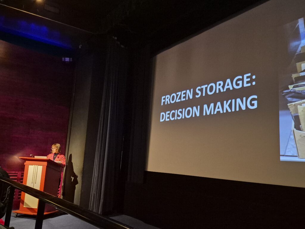 Vanessa speaks from the lectern in the cinema, with the screen showing a slide reading 'Frozen storage: decision making'