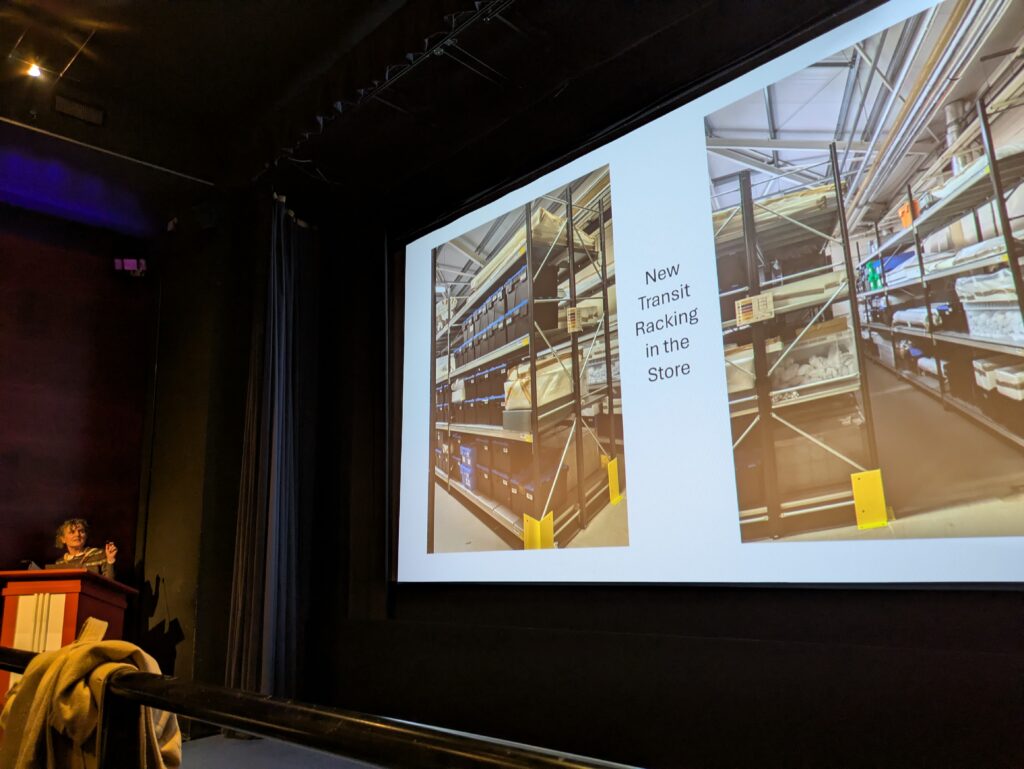 Julie speaks from the lectern, with a slide on screen showing two images of storage racking titled 'New transit racking in the store'