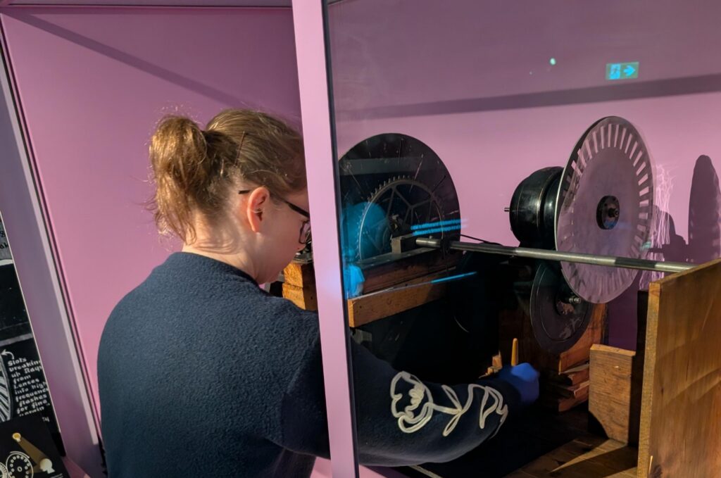 Emma leans into a display case to clean a piece of early broadcast equipment, which is made of wood with two metal discs.