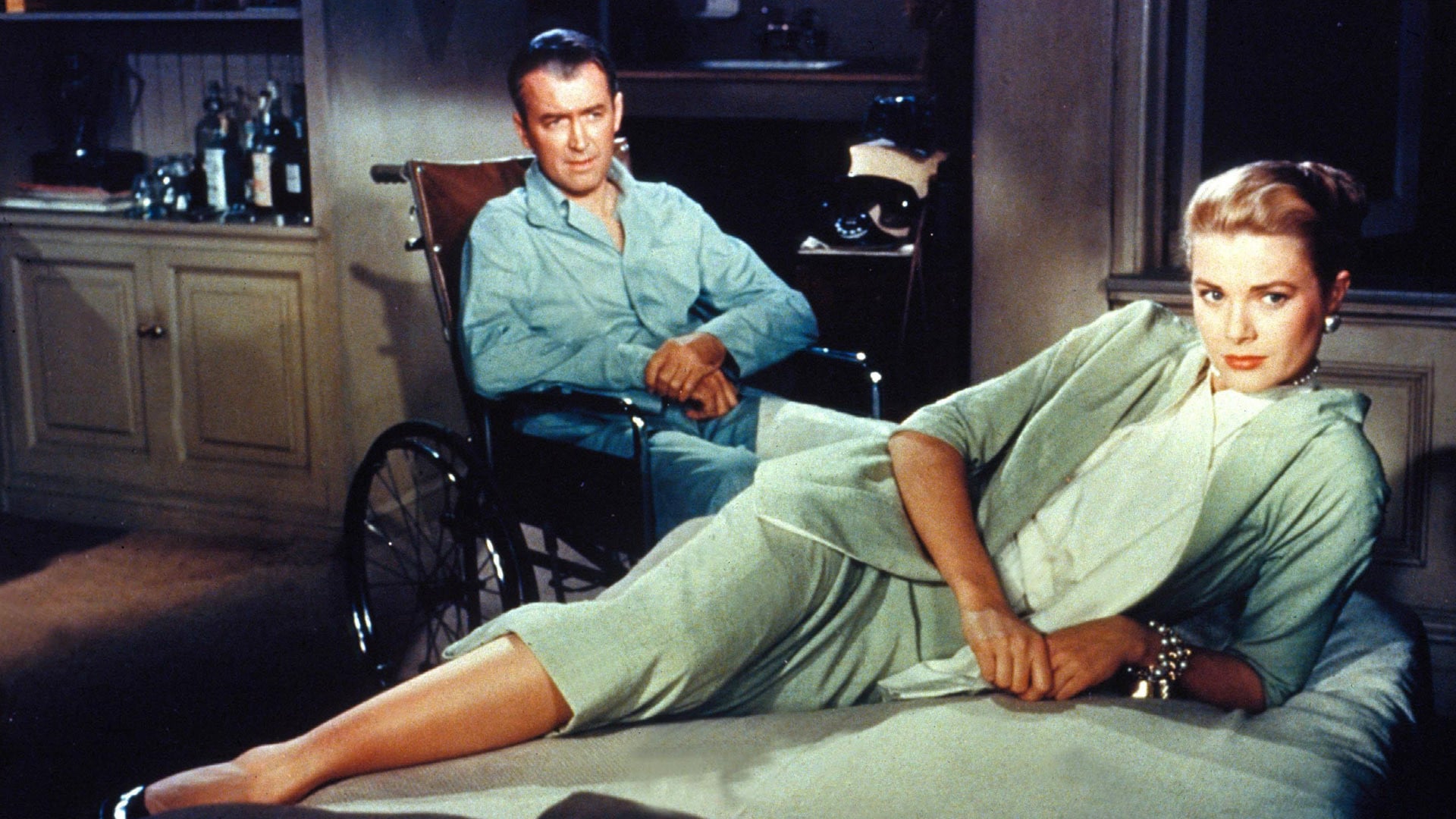 Grace Kelly lays across a sofa wearing a light grey skirt suit, with James Stewart sitting in a wheelchair behind her, wearing blue pyjamas.