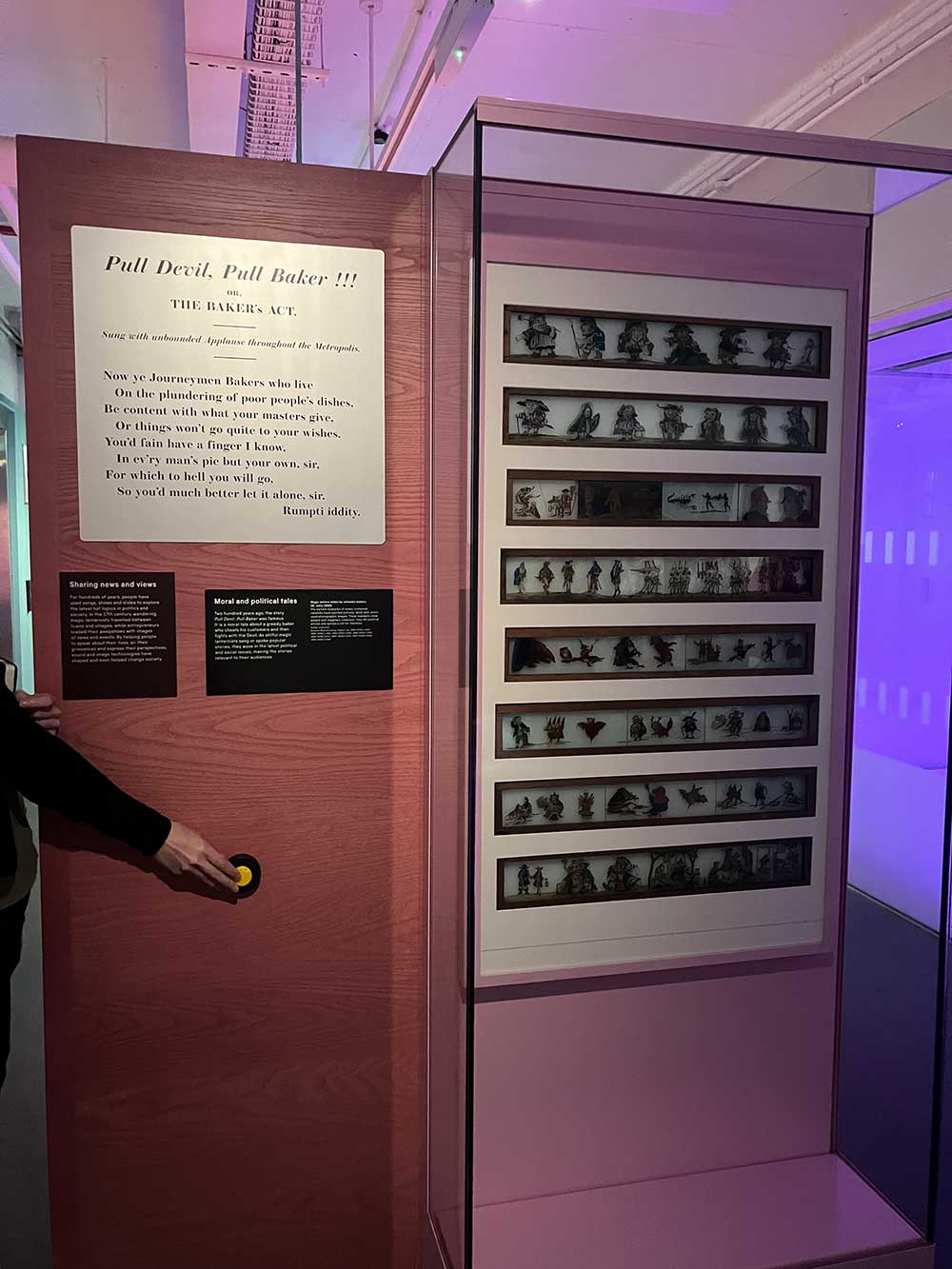 A display of magic lantern slides arranged horizontally in a glass display case. To the right, a hand presses the button to illuminate the slides.