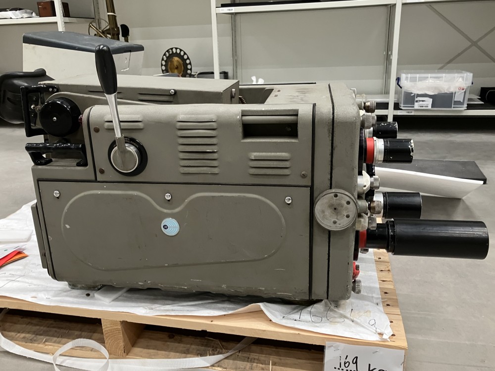 The TV camera seen from the side, with it's handle pointed upwards, on a wooden pallet. Storage racking is in the background