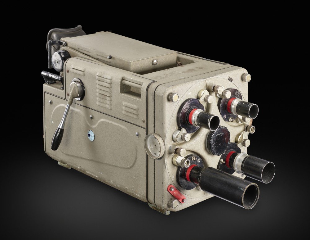 Grey obling cinema camera with 4 cylindrical tubes on the front side, and a silver handle on the side. Set against a black background.