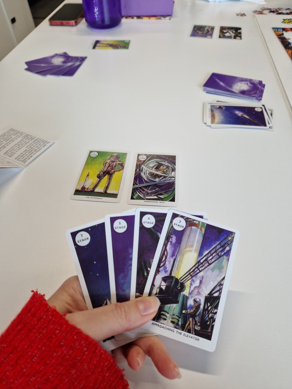 A hand holding four of the game cards, with the game laid out on the table behind.