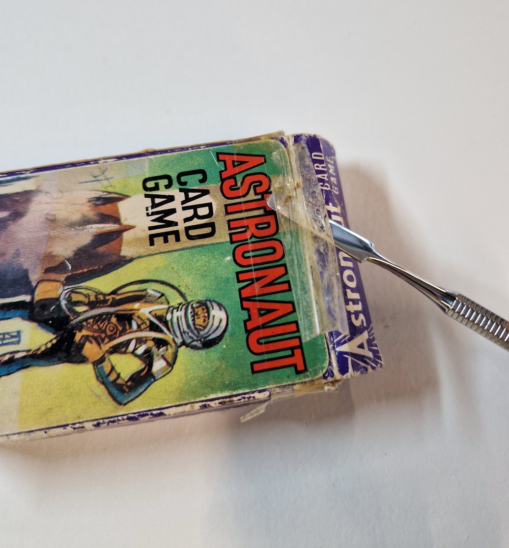 A silver scalpel-like tool being used to lift up the old tape on the card game box.
